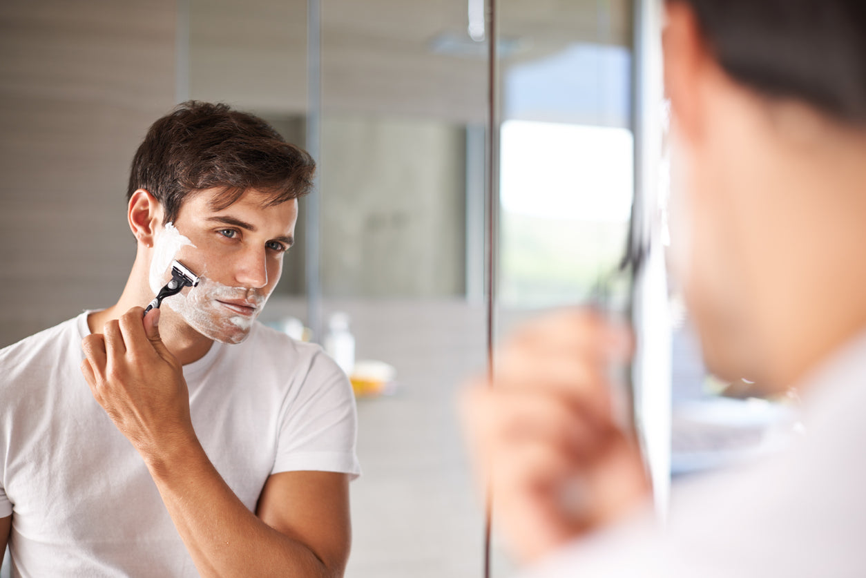 How to Get a Close Shave Without the Pain. Handsome man in white t-shirt is shaving looking in the mirror