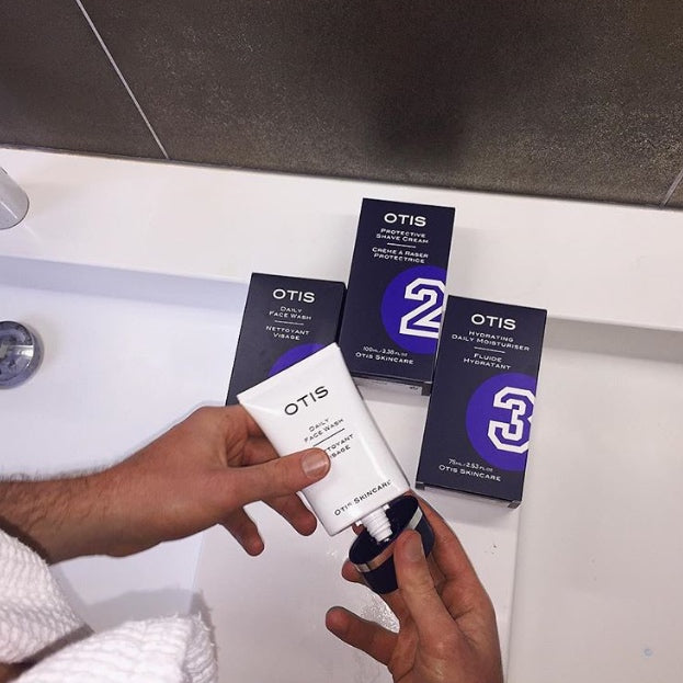 Man taking off lid of tube of OTIS Daily Face Wash at white sink. Other OTIS products in the background