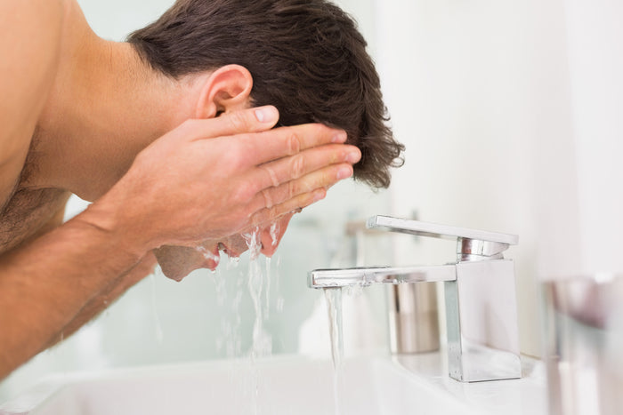 Young man washing with exfoliating face wash - at white sink with shiny chrome taps