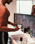 Handsome young man standing at sink opening a Tube of OTIS Protective Shave Cream